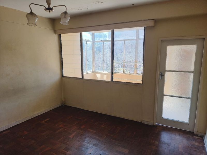 An empty living room featuring original parquet flooring, louvered windows, and a glass panel door, requiring cosmetic updates to the walls and ceiling.