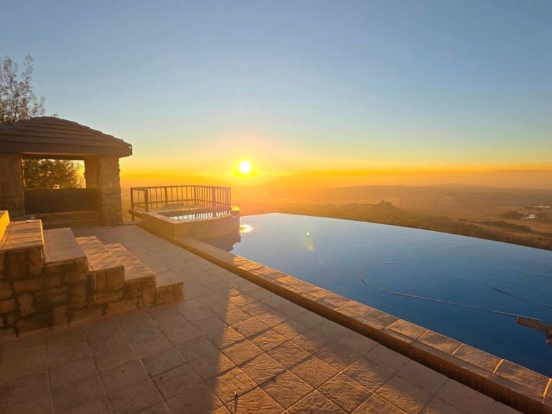 An expansive infinity pool area featuring stone tiling, a covered gazebo, and panoramic sunset views over the surrounding landscape.