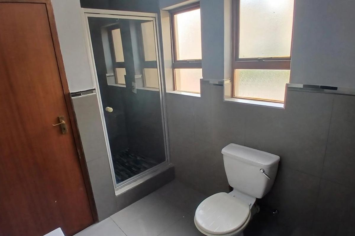 A functional bathroom featuring a glass-enclosed shower, a standard toilet, and neutral grey wall and floor tiling with natural light from casement windows.