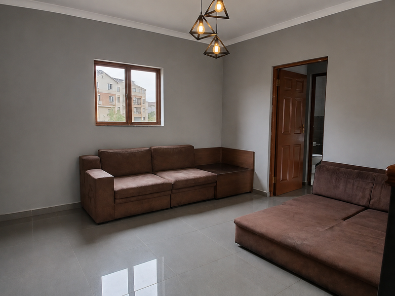 A living room featuring neutral grey walls, large format floor tiles, modern geometric pendant lighting, and a wooden door leading to an adjacent room.
