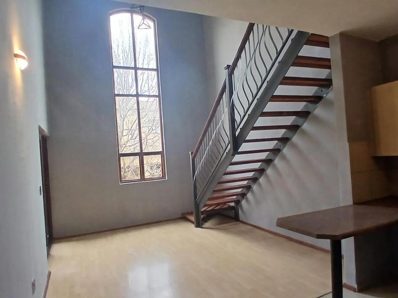 Open plan living area featuring a striking tall arched window, wooden staircase with metal balustrade, and a functional kitchen corner with breakfast bar.