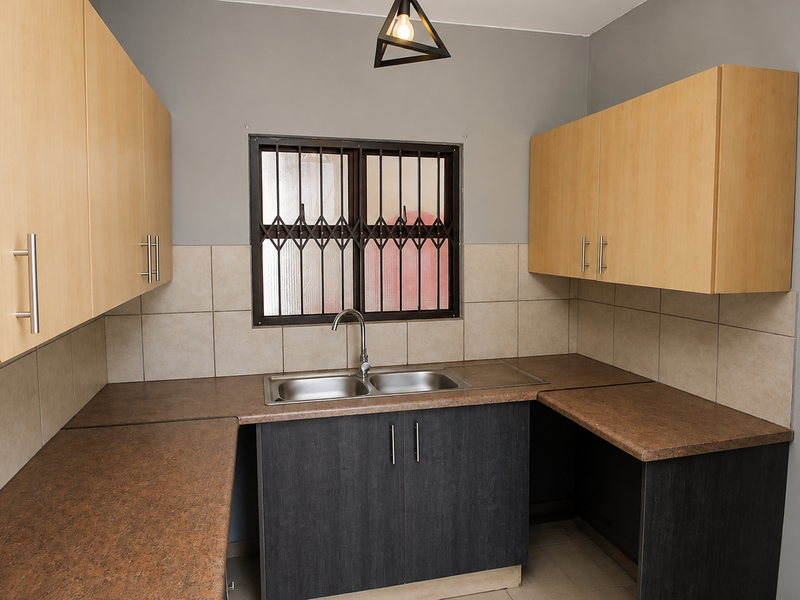 A functional kitchen featuring light wood upper cabinets, dark lower cabinetry, laminate countertops, a double sink, and a tiled backsplash with security window bars.