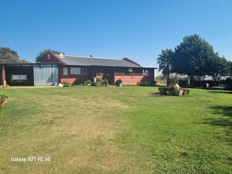 A view of the rear of the farmhouse featuring a large lawn, attached carport, and traditional brick construction.