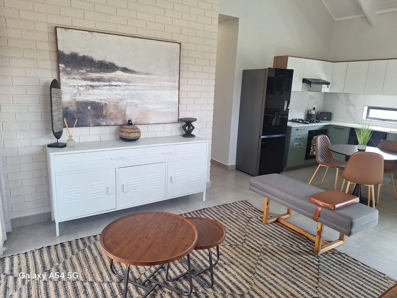 An open-plan living and kitchen area featuring a vaulted ceiling, exposed brick feature wall, modern cabinetry, and tiled flooring throughout.