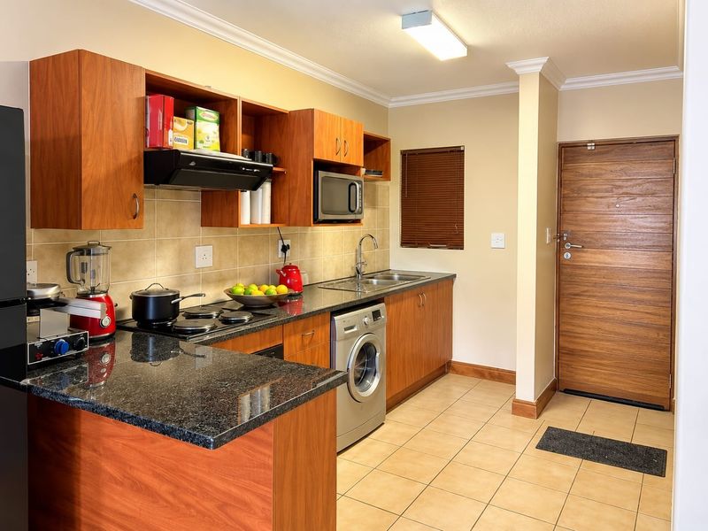 A functional kitchen featuring wood-finish cabinetry, granite countertops, integrated appliances, and tiled flooring, with direct access to the main entrance door.