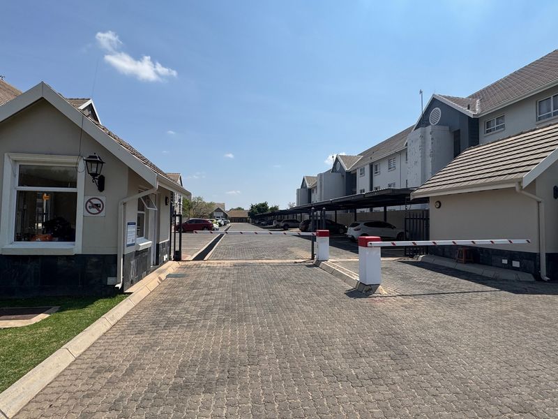 Entrance to the apartment complex featuring a security boom gate, guard house, paved driveway, and covered parking facilities.