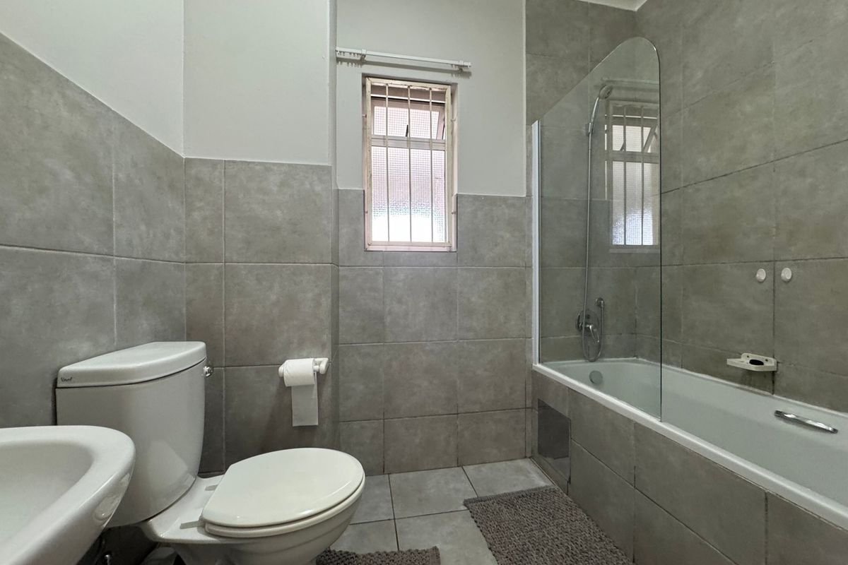 A functional bathroom featuring grey wall and floor tiles, a bathtub with a glass shower screen, a pedestal basin, and a toilet.
