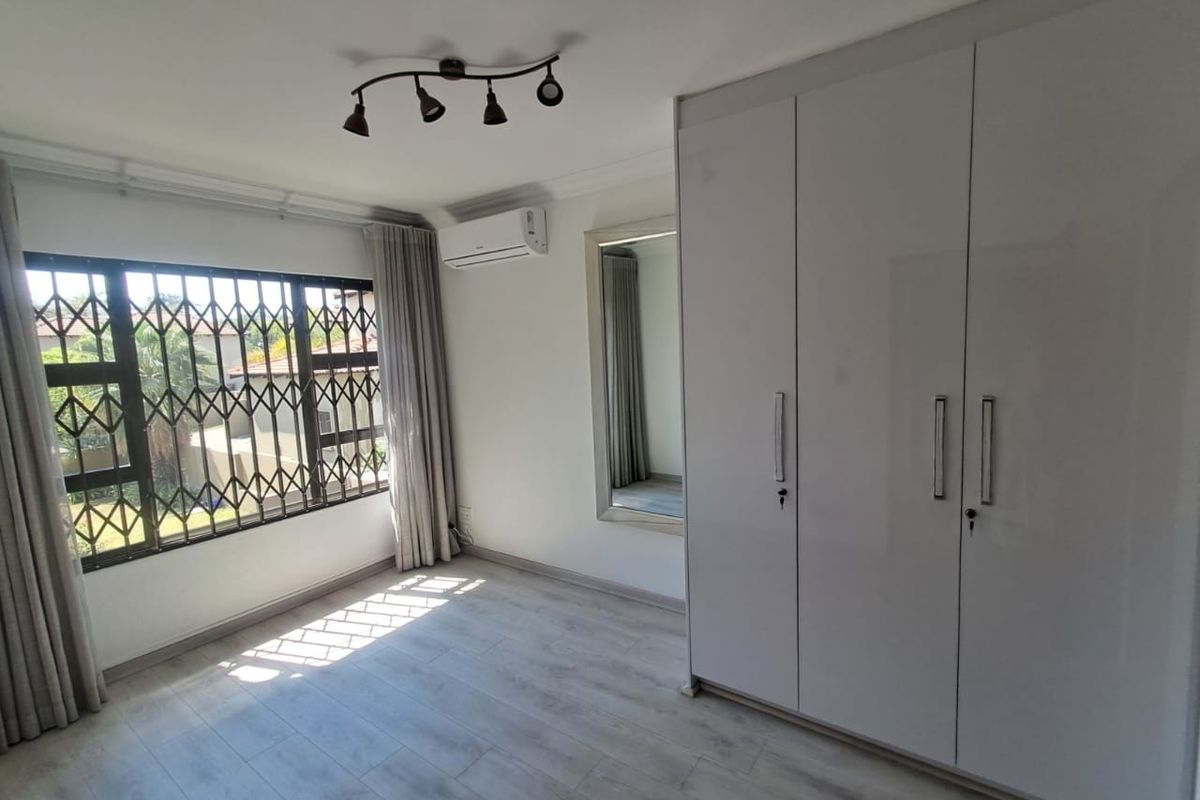 A bright, empty bedroom featuring modern laminate flooring, built-in cupboards, an air conditioning unit, and a security gate on the window.