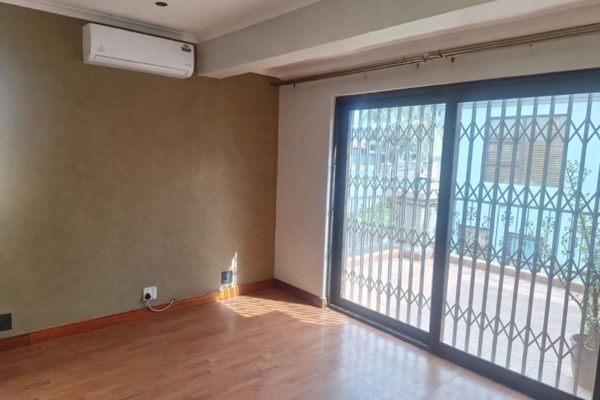An empty living room featuring laminate flooring, a wall-mounted air conditioning unit, and a sliding glass door with a security gate leading outside.