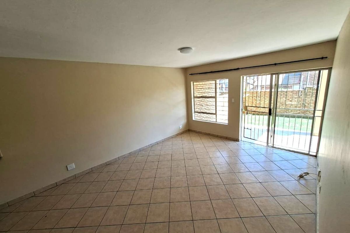 An empty, neutrally painted living room featuring tiled flooring, a sliding glass door with security gate, and large windows allowing natural light.