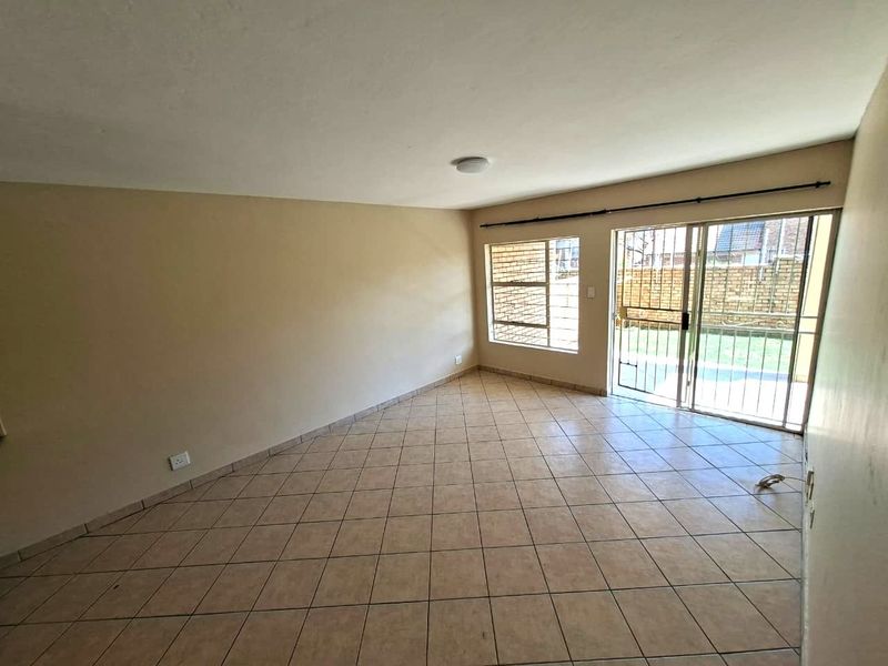 An empty, neutrally painted living room featuring tiled flooring, a sliding glass door with security gate, and large windows allowing natural light.