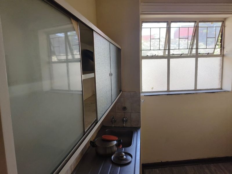 A compact kitchen area featuring a stainless steel sink, wall-mounted storage cabinets, and a tiled splashback, illuminated by a large metal-framed window.
