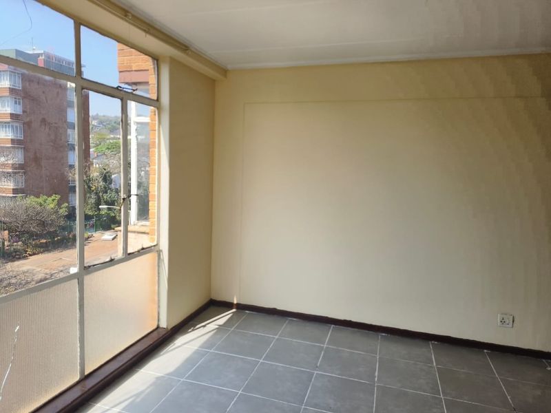 An empty, sunlit living area featuring large metal-framed windows with an urban view and neutral tiled flooring.