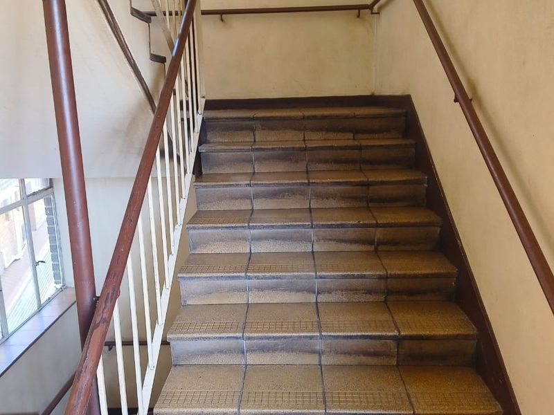 A communal stairwell featuring tiled steps, metal handrails, and a balustrade, showing signs of wear and dated finishes.