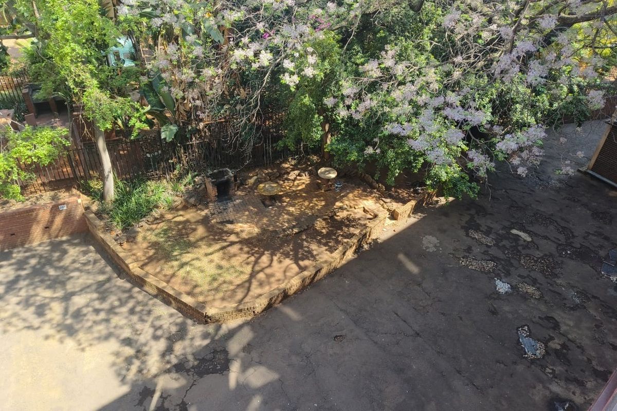 An elevated view of the property grounds featuring a paved driveway, a brick retaining wall, and a built-in outdoor braai area under mature trees.