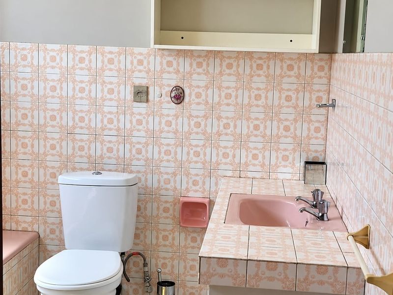 A dated bathroom featuring a pink basin, tiled vanity, and wall-mounted shelving. The space requires modernization of finishes and fixtures.