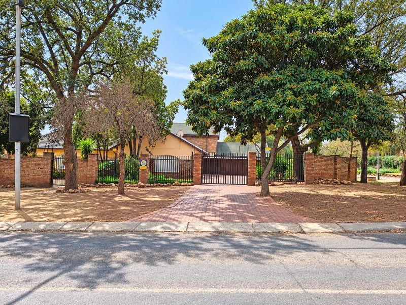 Front view of the property featuring a paved driveway, secure metal gate, and brick boundary wall surrounded by mature trees.