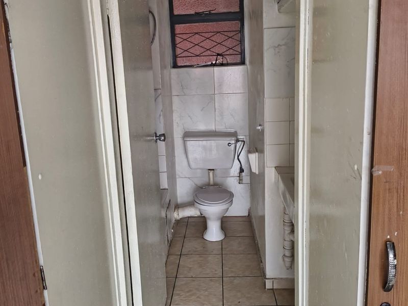 A compact, dated bathroom featuring tiled flooring, wall tiles, a standard toilet, and a small basin with a window providing natural light.