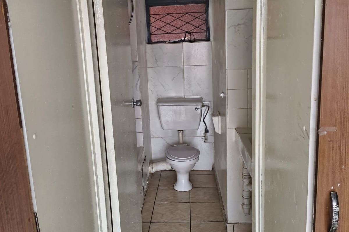 A compact, dated bathroom featuring tiled flooring, wall tiles, a standard toilet, and a small basin with a window providing natural light.
