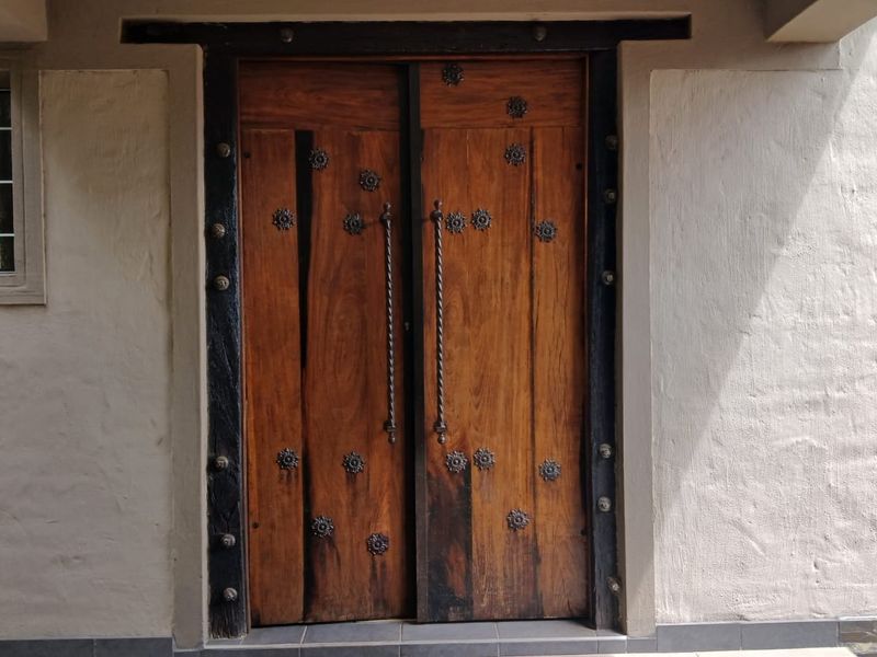 A rustic entrance featuring solid wood double doors with decorative metal detailing, set within a textured plastered wall and adjacent grid window.