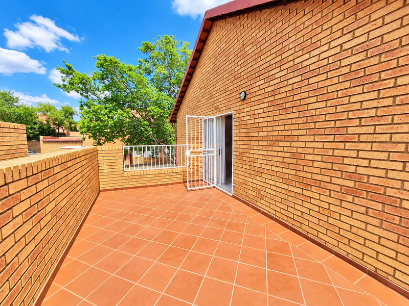 A spacious outdoor balcony featuring terracotta-style tiled flooring, brick walls, and a secure metal gate entrance.