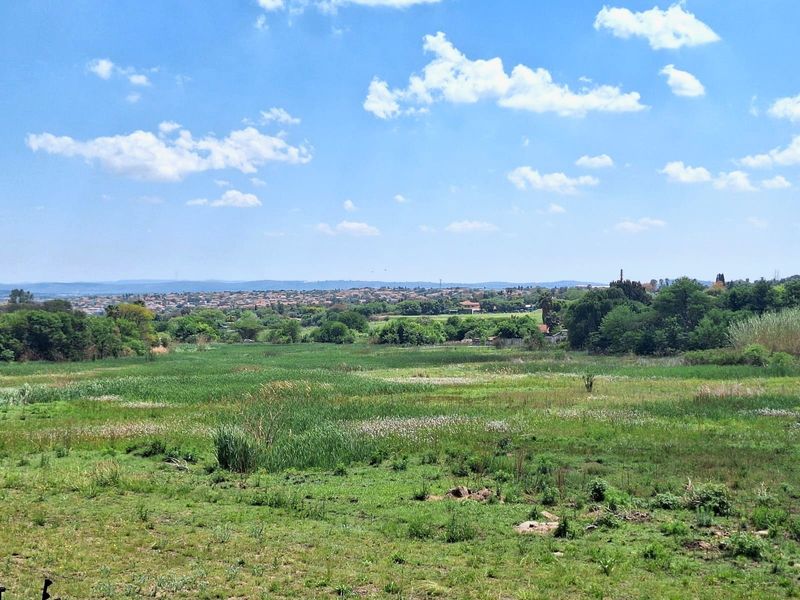 A scenic landscape view overlooking a green field with distant residential properties under a clear blue sky.