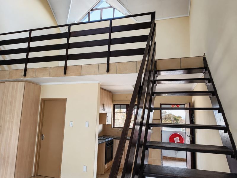 Open-plan living area featuring a mezzanine level, timber staircase, and a functional kitchen with built-in cabinetry and appliances under a vaulted ceiling.