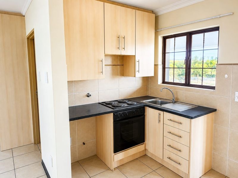 A compact, functional kitchen featuring light wood-finish cabinetry, black countertops, a built-in oven and hob, and neutral tiled finishes.