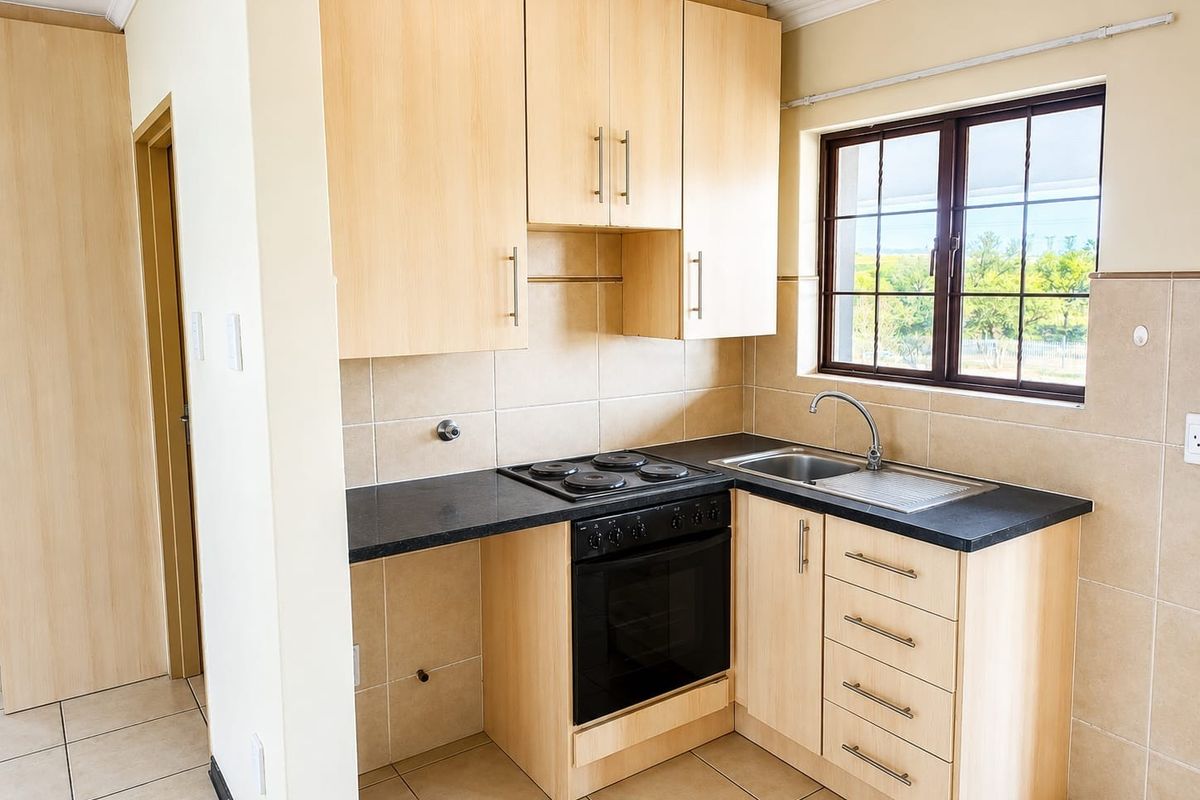 A compact, functional kitchen featuring light wood-finish cabinetry, black countertops, a built-in oven and hob, and neutral tiled finishes.