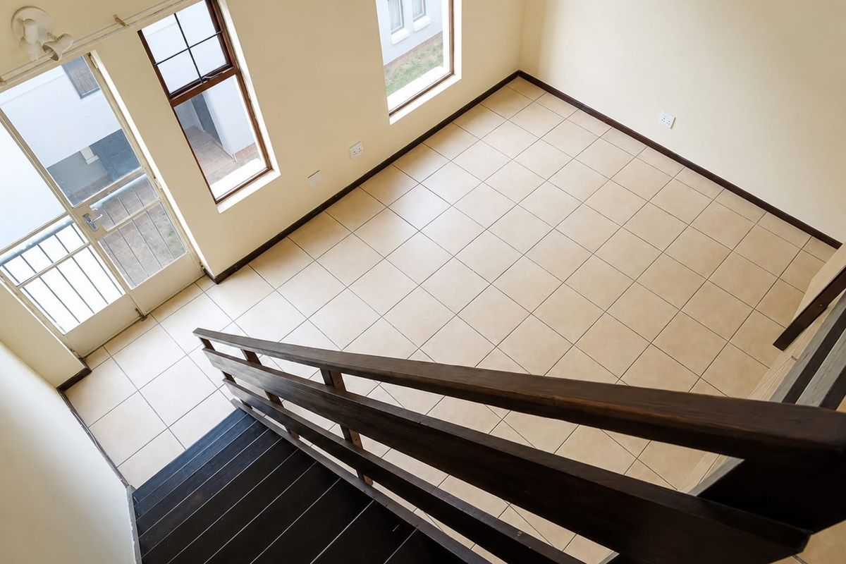 An empty, well-lit living area featuring neutral tiled flooring, a wooden staircase with a matching handrail, and access to an outdoor balcony.