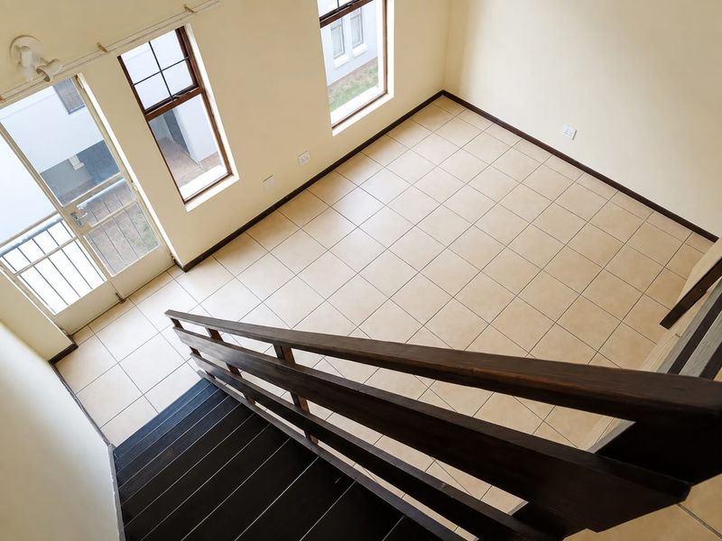 An empty, well-lit living area featuring neutral tiled flooring, a wooden staircase with a matching handrail, and access to an outdoor balcony.
