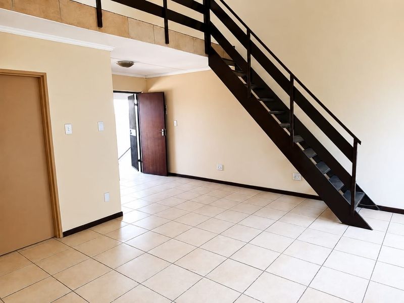 An open-plan living area featuring tiled flooring, a dark timber staircase leading to a mezzanine level, and a secure entrance door.
