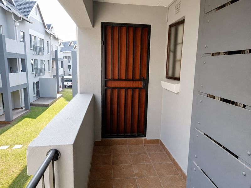 A clean, covered balcony area featuring tiled flooring, a secure metal gate, and a view overlooking the apartment complex grounds.