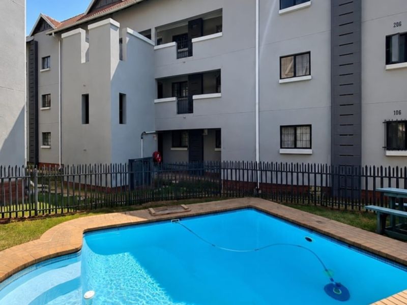 A well-maintained communal swimming pool area featuring brick coping and metal perimeter fencing, set against the backdrop of a modern apartment building complex.
