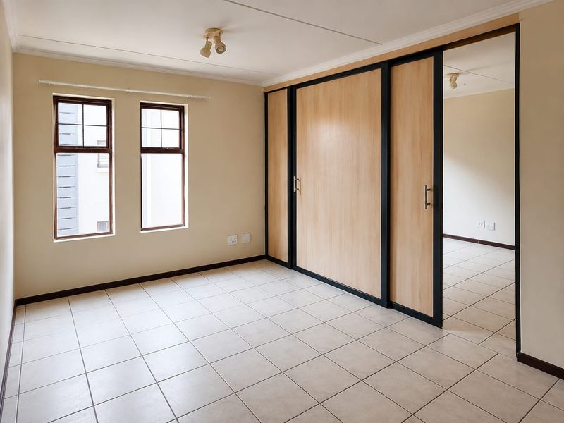 A bright, empty bedroom featuring neutral tiled flooring, built-in sliding door wardrobes, and natural light from dual casement windows.