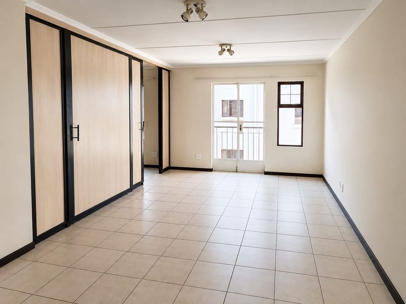 A bright, empty bedroom featuring neutral tiled flooring, built-in cupboards with modern trim, and a sliding door leading to a small balcony.