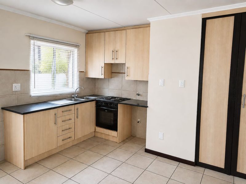 A clean, functional kitchen featuring light wood-finish cabinetry, black countertops, built-in oven and hob, tiled backsplash, and neutral floor tiles.