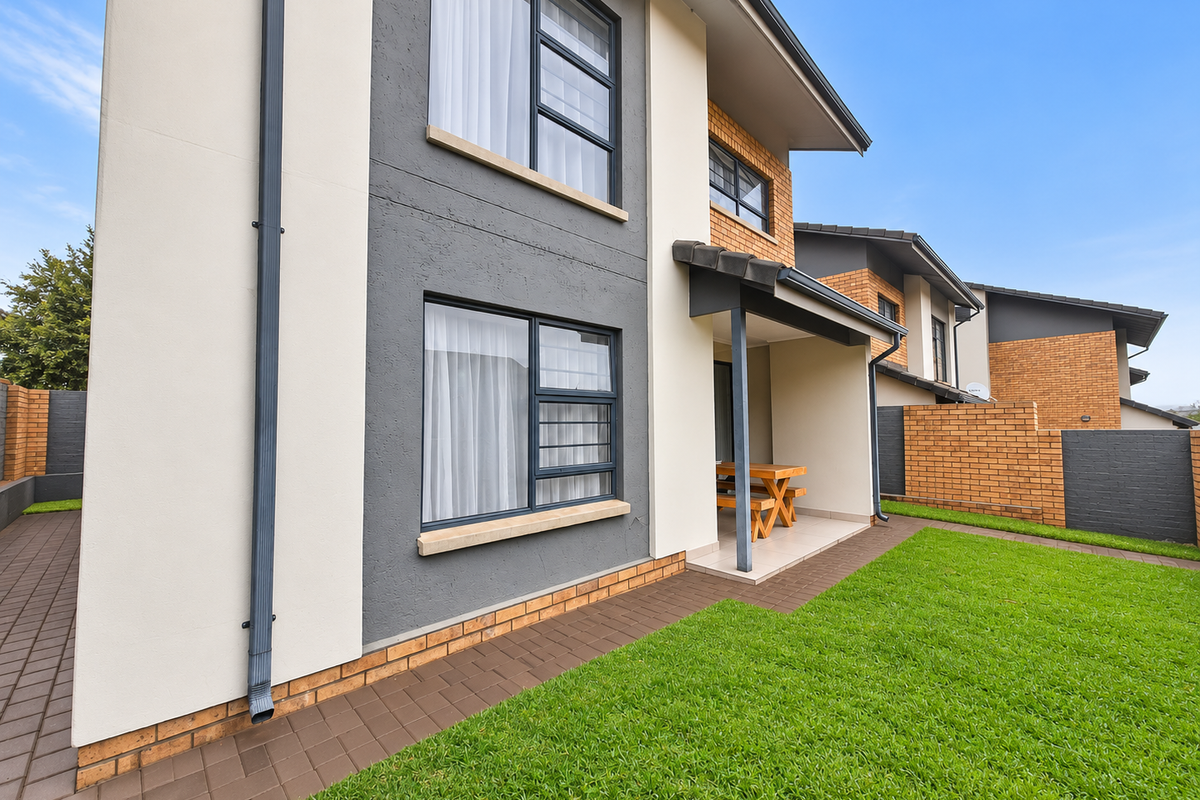 Rear view of a modern townhouse featuring a covered patio, manicured lawn, paved pathways, and a combination of brick and rendered exterior finishes.