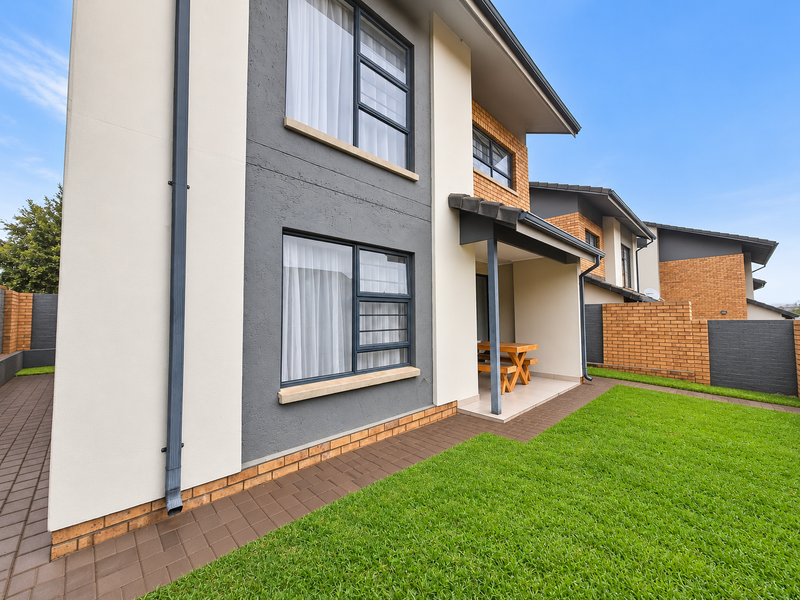 Rear view of a modern townhouse featuring a covered patio, manicured lawn, paved pathways, and a combination of brick and rendered exterior finishes.