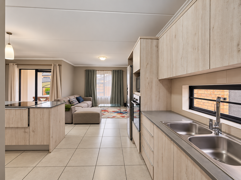 Modern open-plan kitchen and living area featuring light wood-grain cabinetry, tiled floors, and seamless flow to the outdoor patio through sliding glass doors.