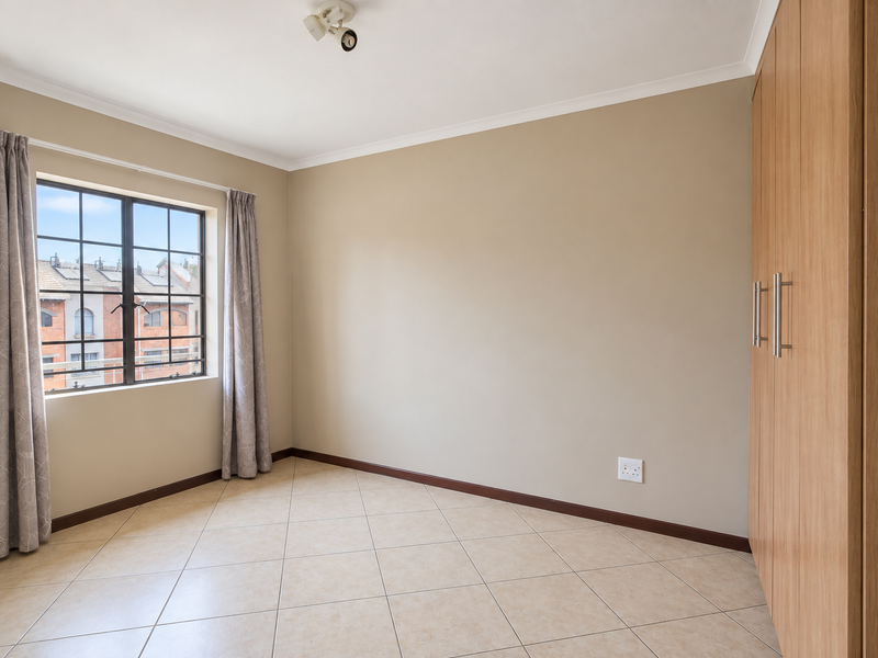 A bright, empty bedroom featuring neutral walls, tiled flooring, and built-in wooden cupboards, offering a clean and versatile space.