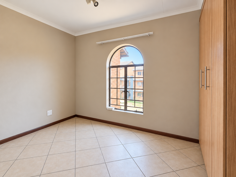 A bright, empty bedroom featuring neutral walls, tiled flooring, and a built-in wooden wardrobe with an arched window providing natural light.