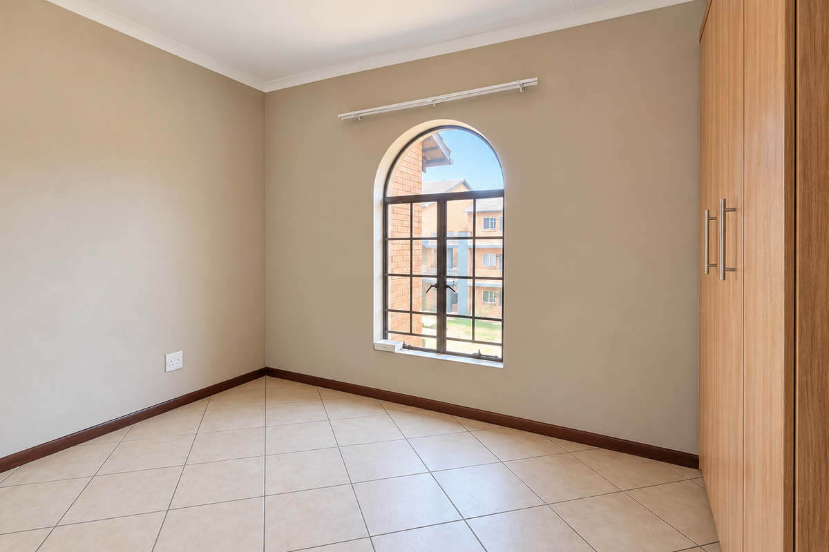 A bright, empty bedroom featuring neutral walls, tiled flooring, and a built-in wooden wardrobe with an arched window providing natural light.