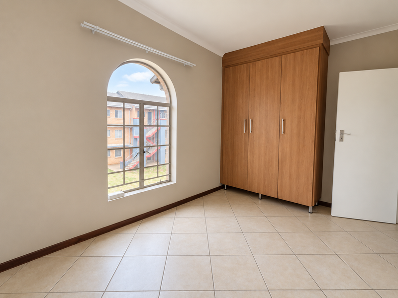 A bright, empty bedroom featuring neutral tiled flooring, a large arched window, and a built-in wooden wardrobe.