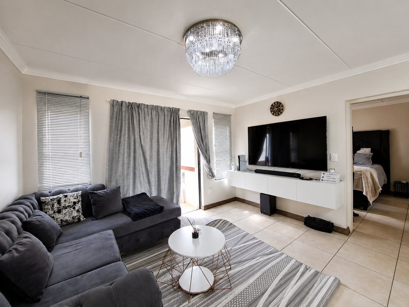 A well-maintained living room featuring tiled flooring, a modern wall-mounted media console, and a decorative crystal chandelier, with direct access to a balcony.
