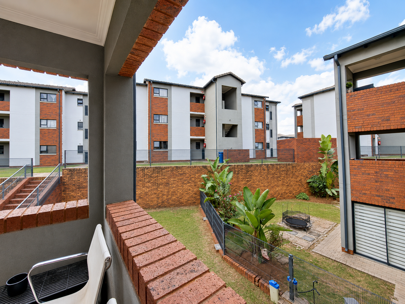 A view from a covered balcony overlooking a communal garden area and brick-finished apartment complex buildings.