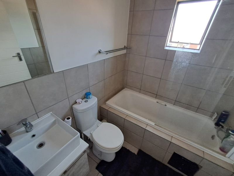 A functional bathroom featuring a built-in bathtub with a glass screen, a wall-mounted basin, and a toilet, finished with neutral wall and floor tiling.