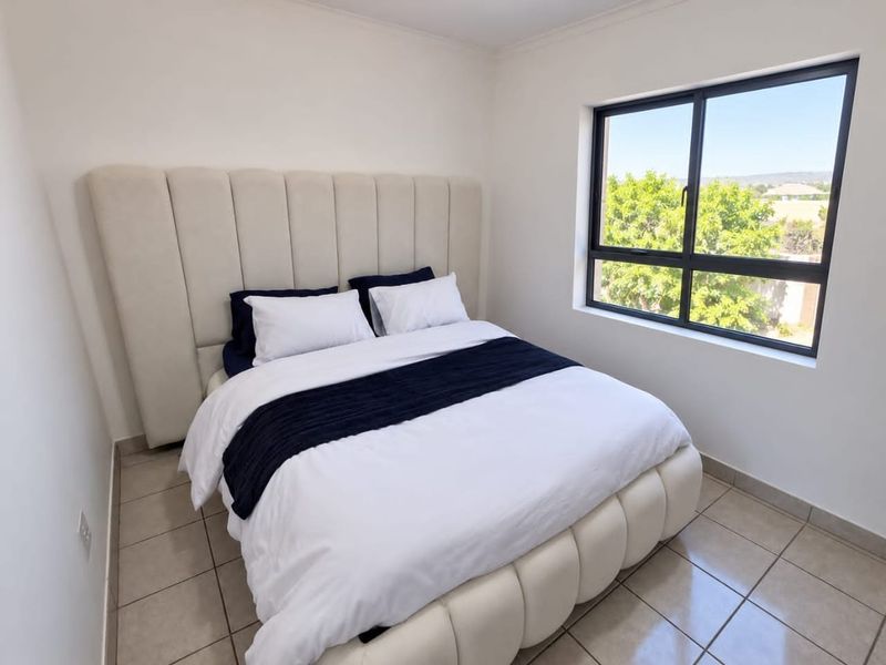 A bright bedroom featuring neutral tiled flooring and a large window with an aluminium frame, offering natural light and a pleasant outdoor view.