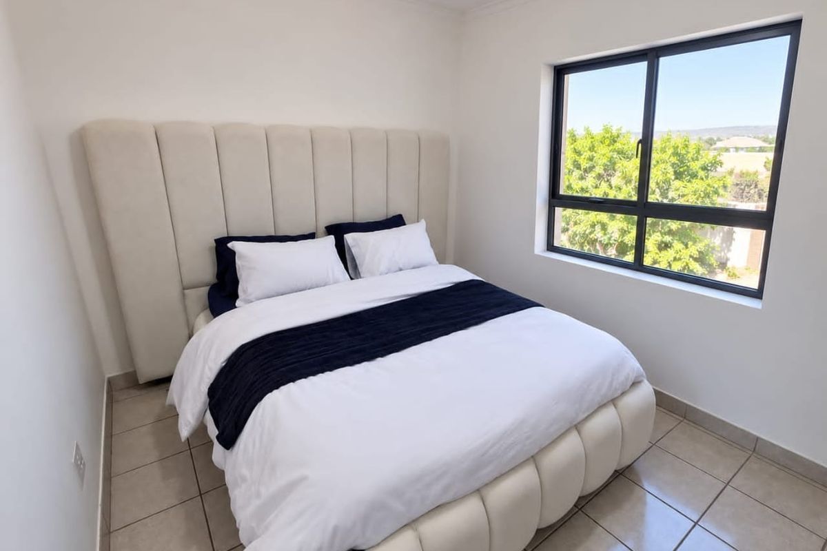 A bright bedroom featuring neutral tiled flooring and a large window with an aluminium frame, offering natural light and a pleasant outdoor view.