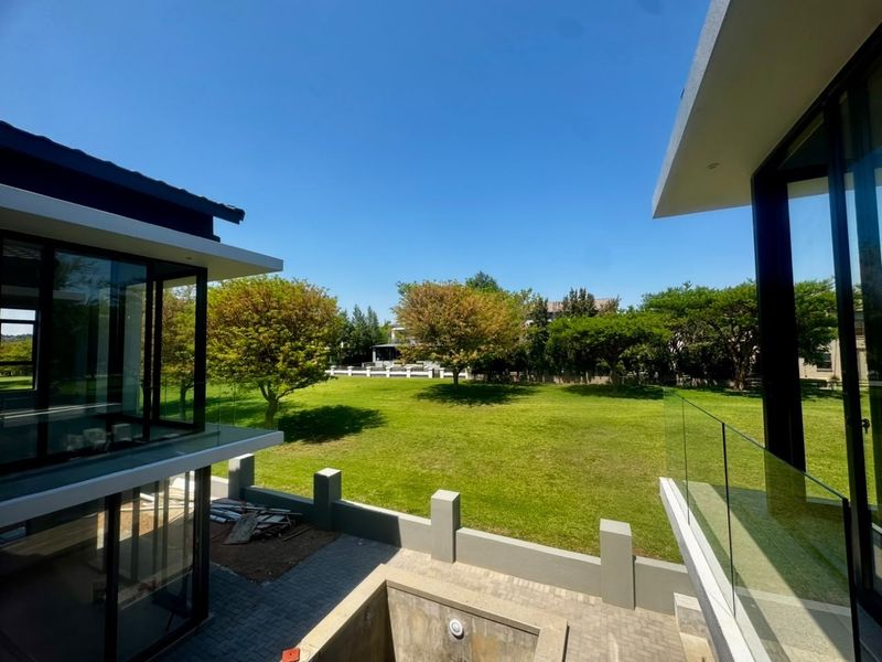 Rear view of a modern property featuring glass balustrades, a paved patio area, and an unfinished swimming pool overlooking a large garden.